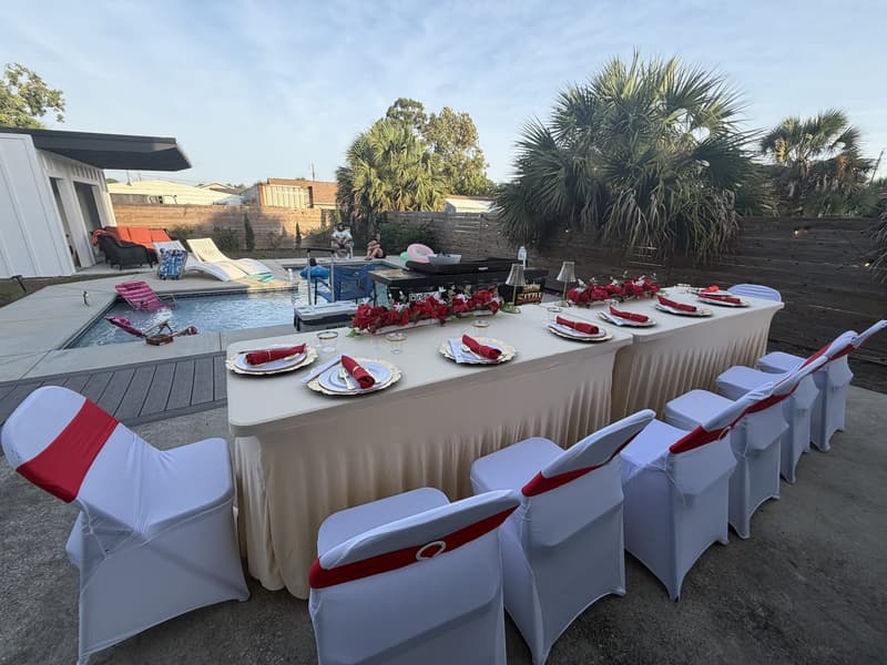 A long table dressed in white with red sashes and roses beside a pool, palms and low resort buildings behind it