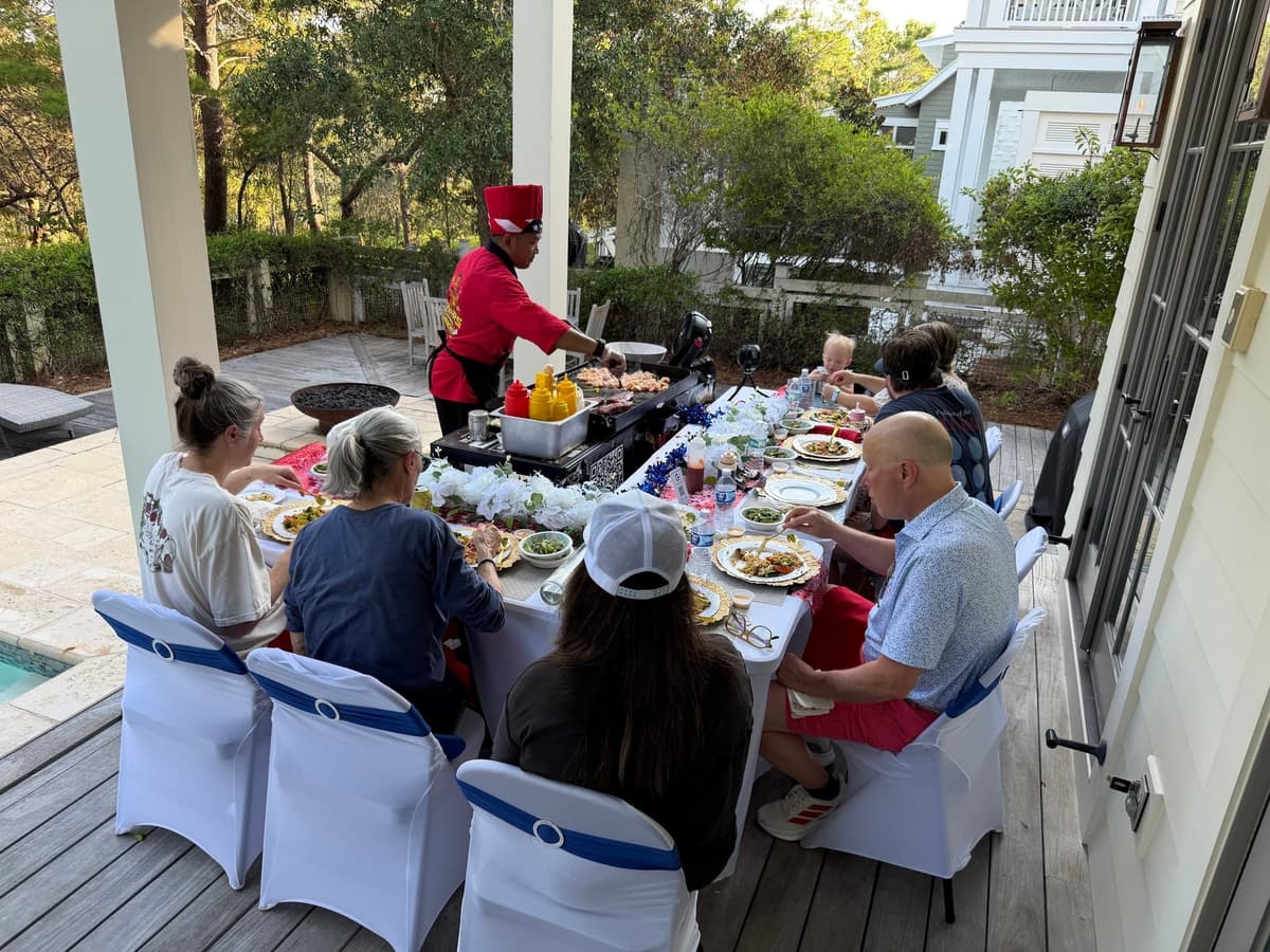 Chef cooking at the flat-top set at the head of a long table of seated guests on a shaded deck
