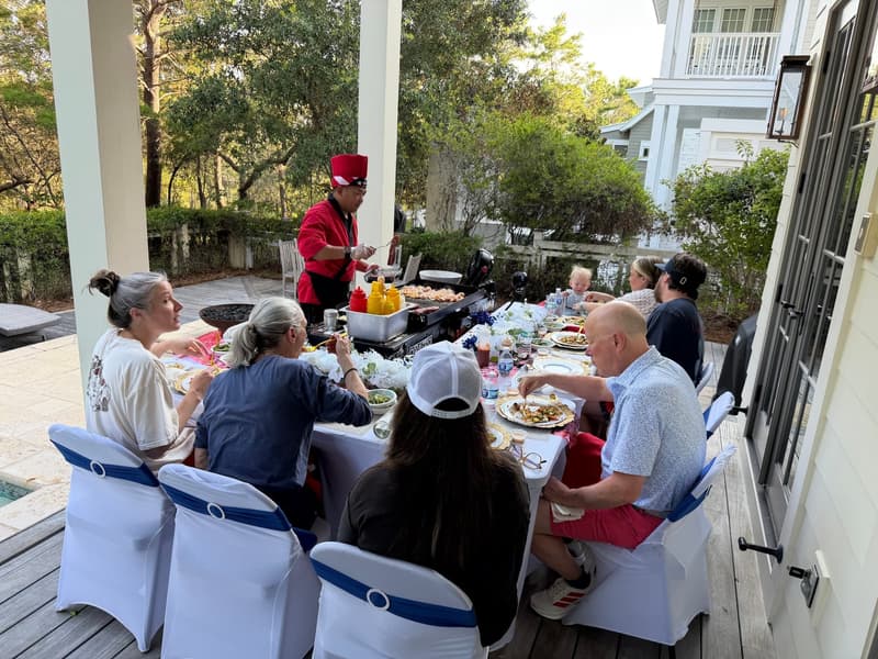 Guests seated the length of a long table on a shaded porch while the chef works the flat-top at the head