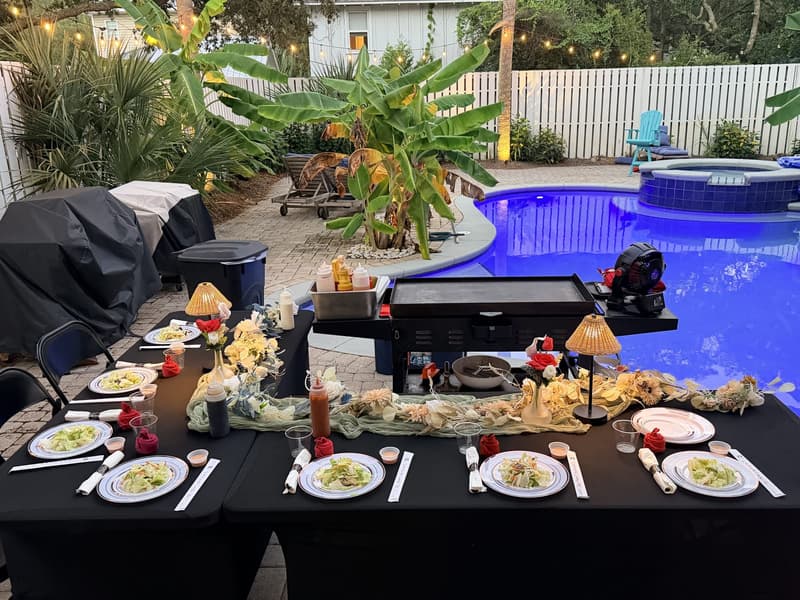 A long table set beside a lit swimming pool at dusk, the flat-top grill standing ready at one end