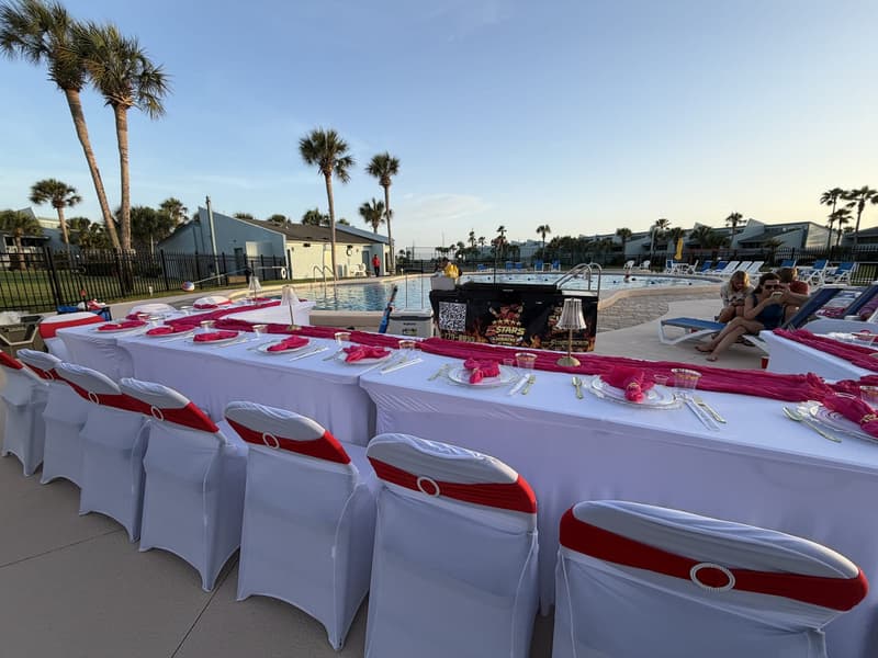 Long tables in white covers with red sashes set out beside a pool at sunset, palms and low buildings behind