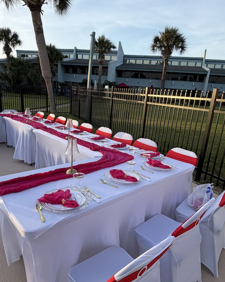Long table dressed with a deep pink runner and gold place settings beside a pool, dark railing behind